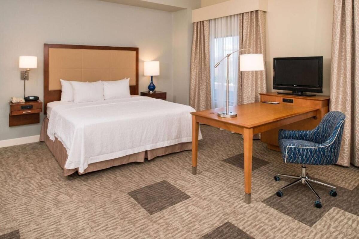 Quarto de hotel com uma cama, uma escrivaninha, uma cadeira, uma televisão e duas luminárias de cabeceira.