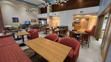 Uma área de refeições moderna, com várias mesas e cadeiras, uma televisão e um balcão com o menu.