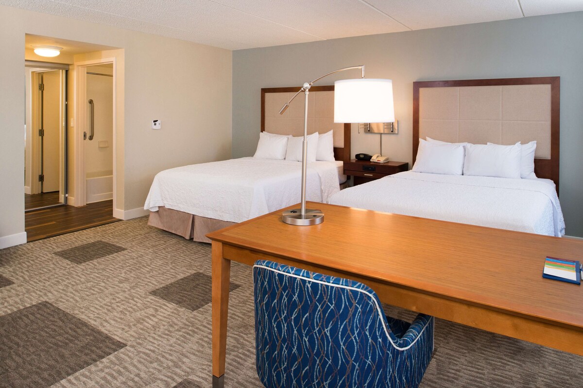 A hotel room with two beds, a desk, a chair, a lamp, and a nightstand.