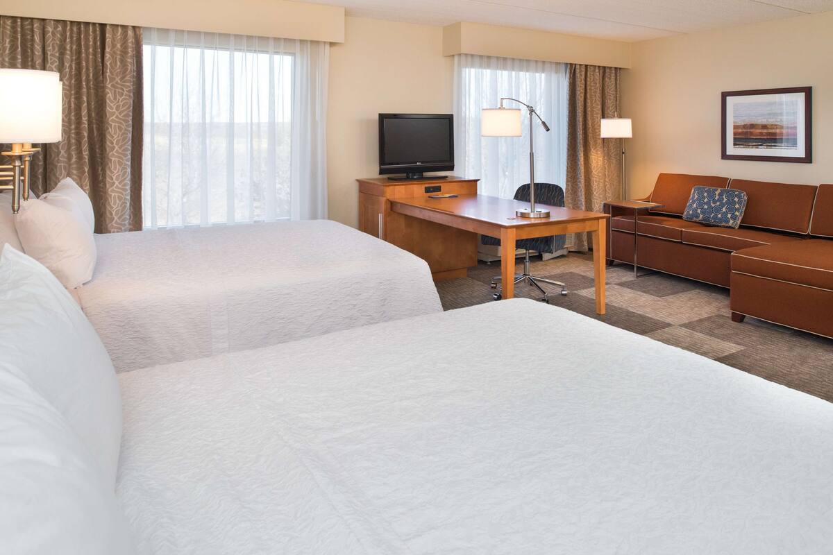 A hotel room with two beds, a desk with a television, a sofa, and a lamp.