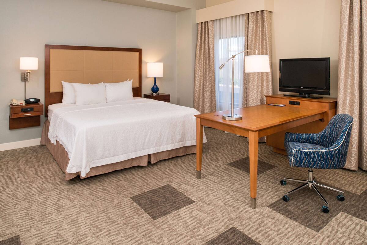 A hotel room with a bed, bedside tables, a desk with a chair, a television, and a window with curtains.