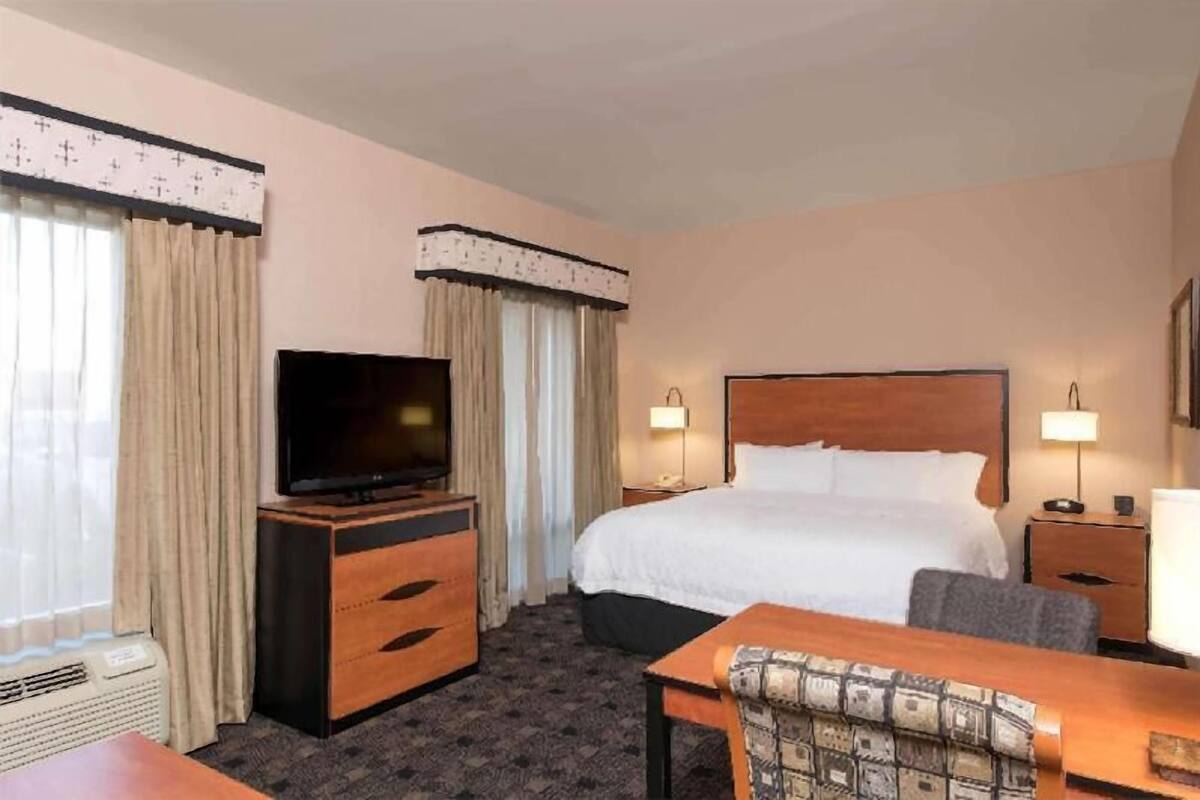 A hotel room with a bed, a TV on a wooden dresser, a desk with a chair, and two bedside lamps.
