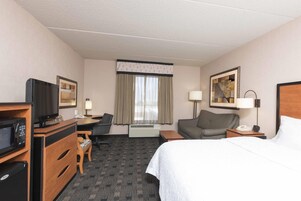 A hotel room with a bed, a desk, a chair, a television, a microwave, a lamp, and a framed picture on the wall.