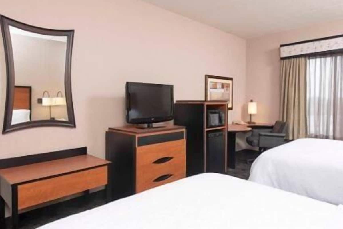 A hotel room with a bed, a TV on a wooden stand, a desk with a chair, a mirror, and a window with curtains.
