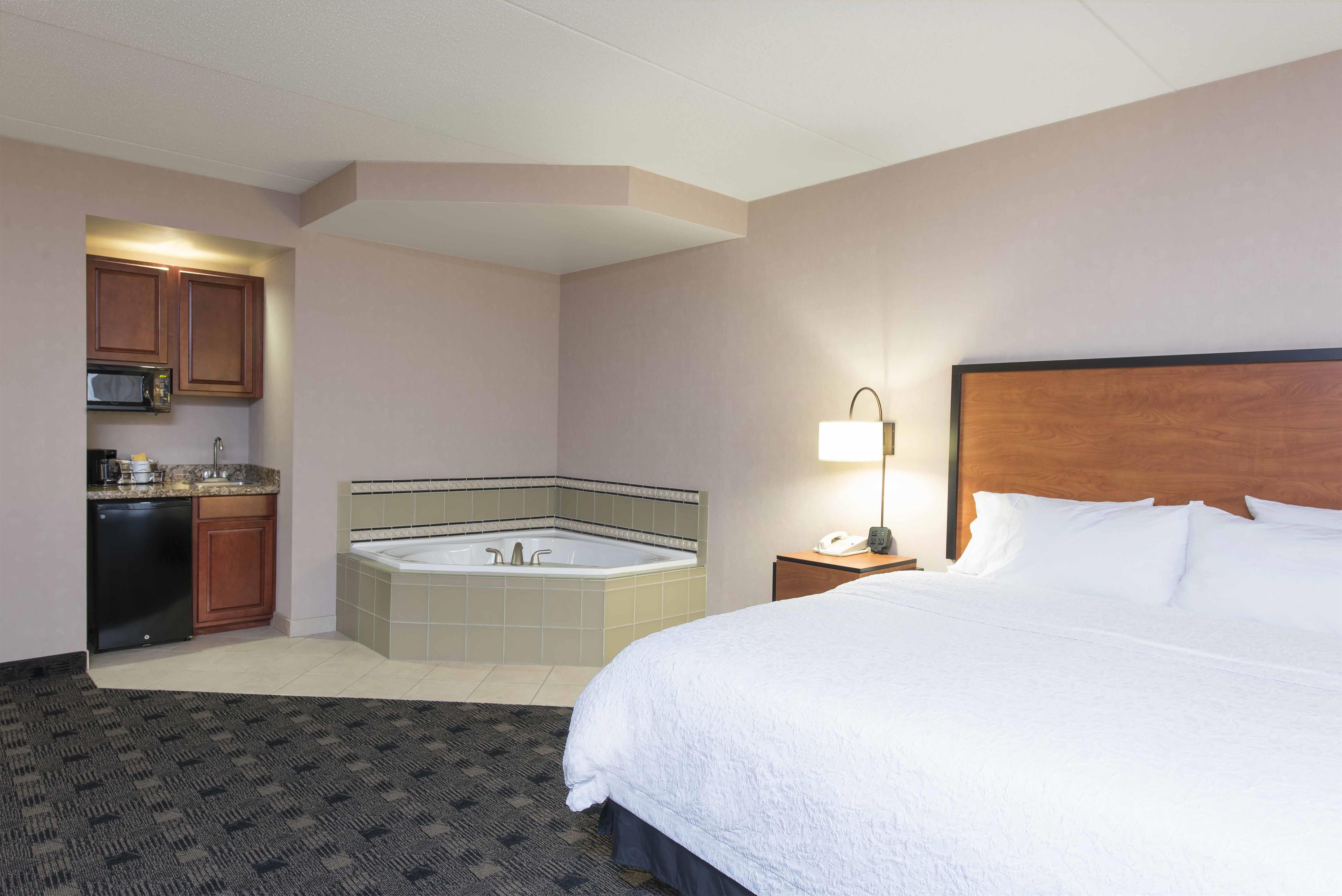 Habitación de hotel con cama, baño con bañera, kitchenette y mesita de noche con lámpara.