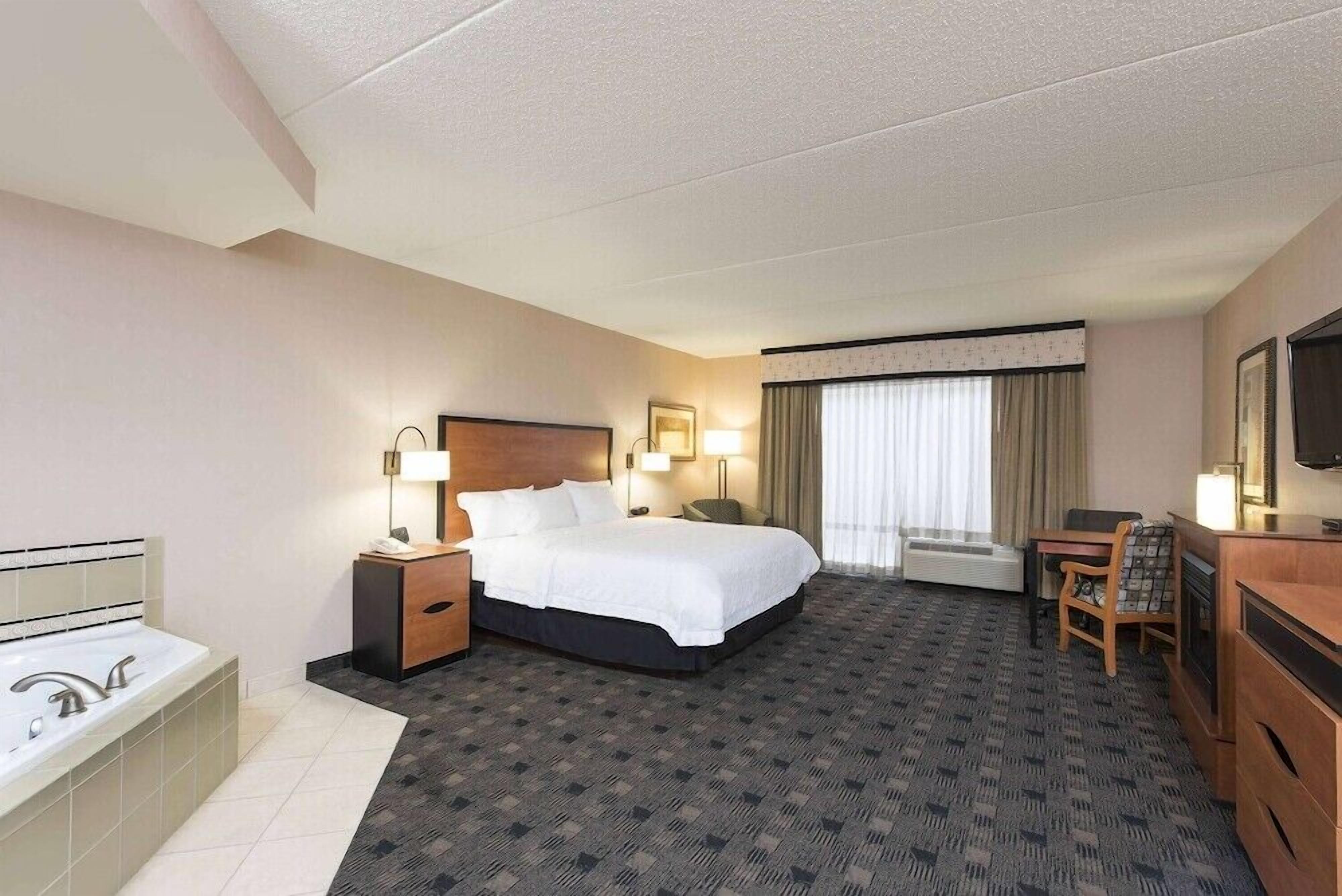 Ein Hotelzimmer mit Bett, Schreibtisch, Sessel, Fernseher, Lampe, Nachttisch und einem Badezimmer mit Badewanne.