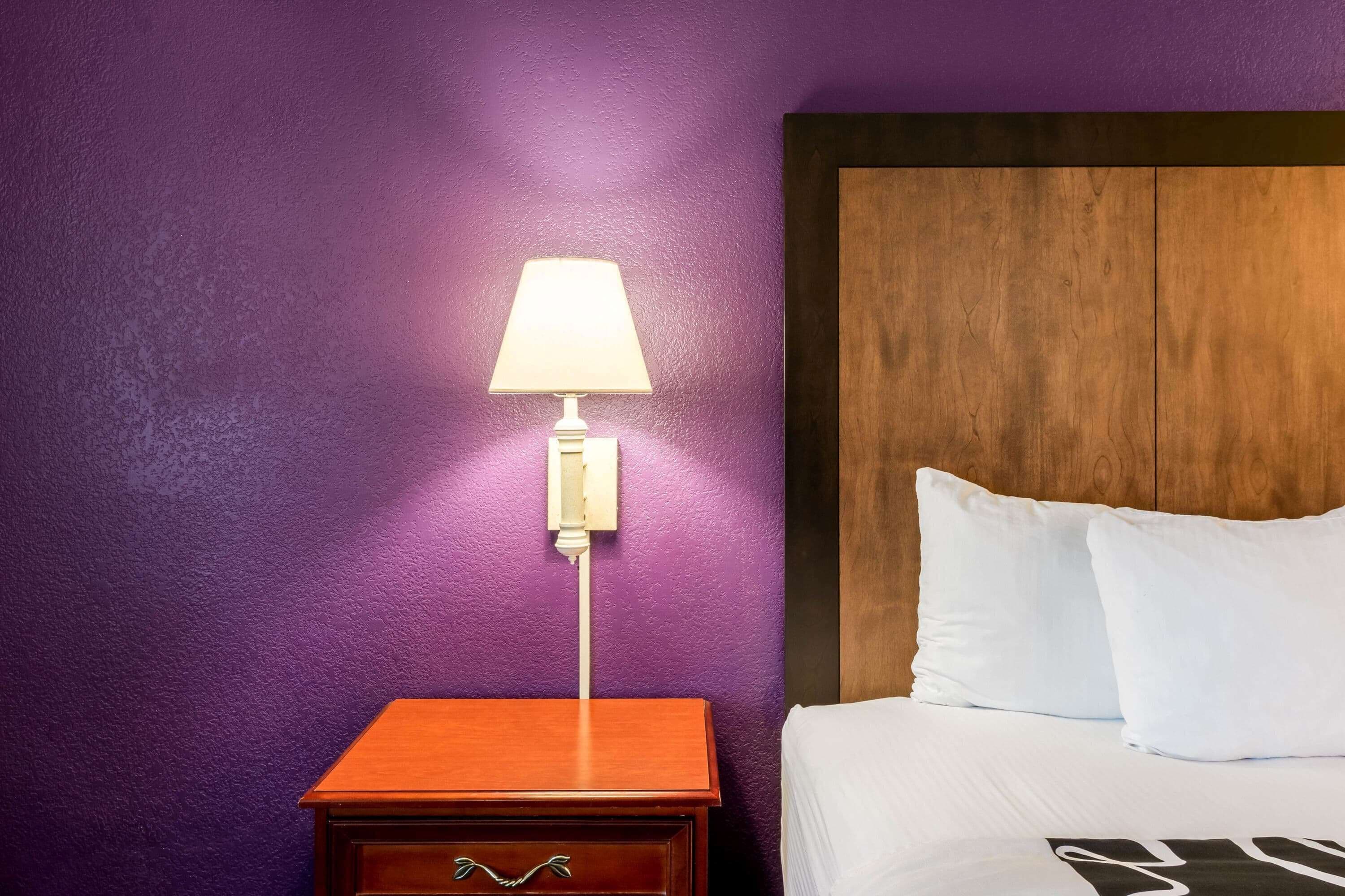 Een hotelkamer met een houten nachtkastje, een wandlamp en een bed met witte lakens.
