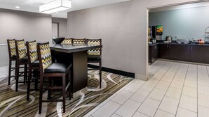 Un comedor moderno con una mesa rectangular y cuatro sillas, cada una con un cojín estampado. Se ve un área de cocina a la derecha con un refrigerador y una cafetera.