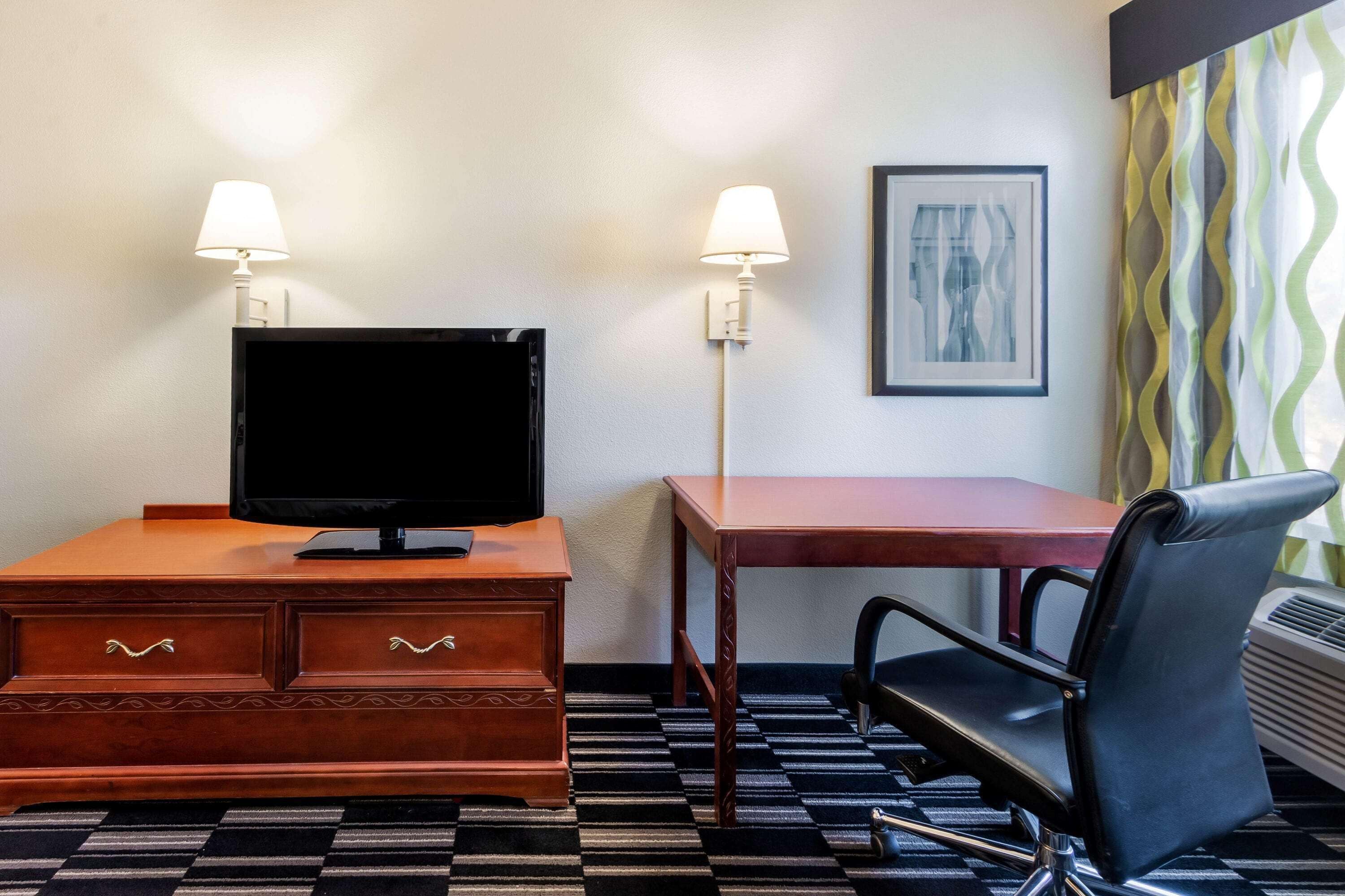 A hotel room with a wooden desk, a television, a chair, and two wall-mounted lamps.