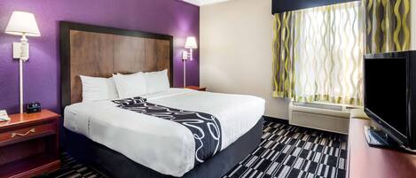 A hotel room with a large bed, a flat-screen TV, a nightstand with a lamp, and a window with patterned curtains.