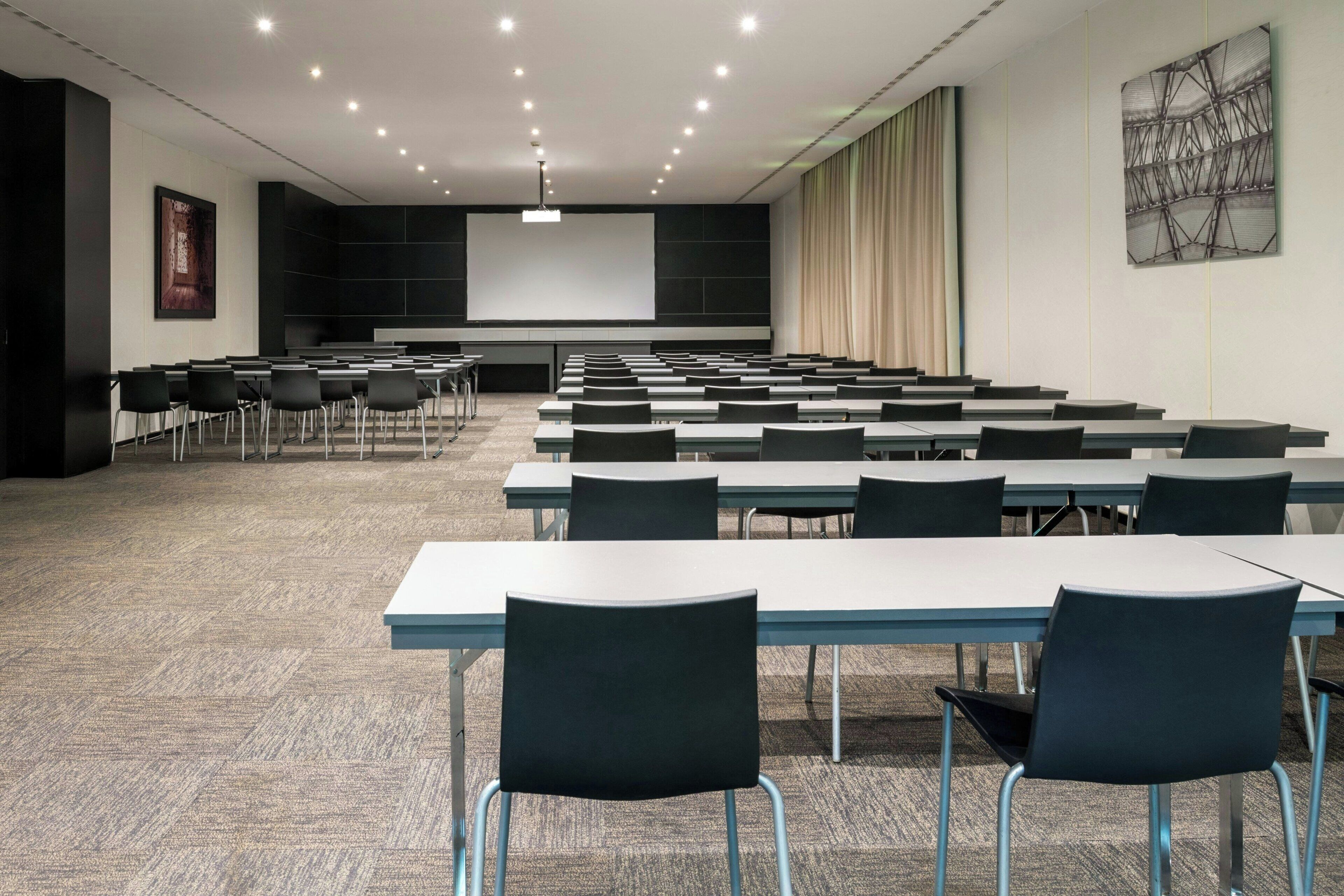Un salón de conferencias con filas de mesas y sillas, una pantalla de proyector y un cuadro grande en la pared.