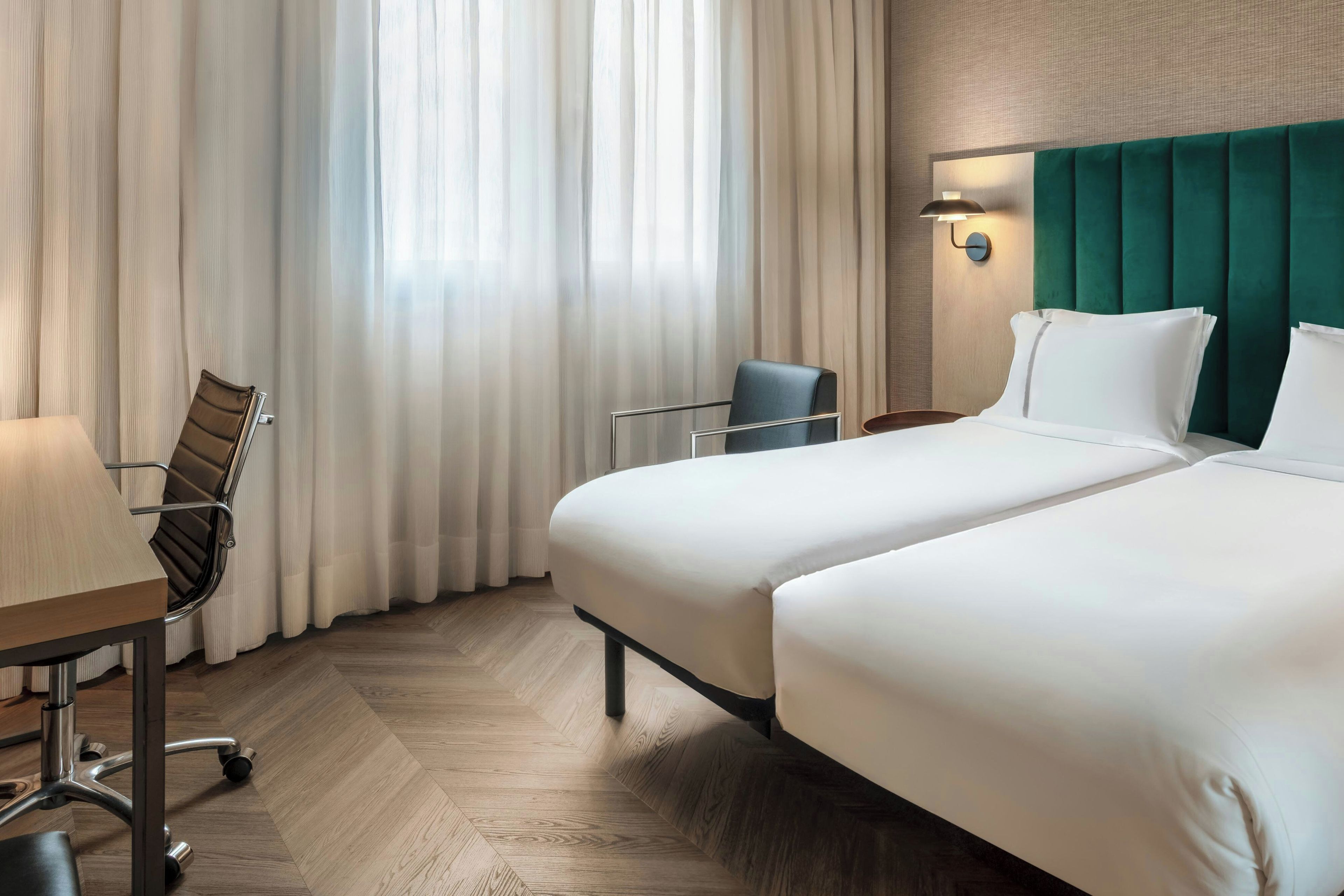 A hotel room with two beds, a desk with a chair, a lamp, and a window with curtains.