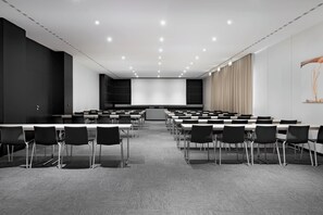 Una sala conferenze con file di tavoli e sedie, un podio e un grande schermo di fronte.