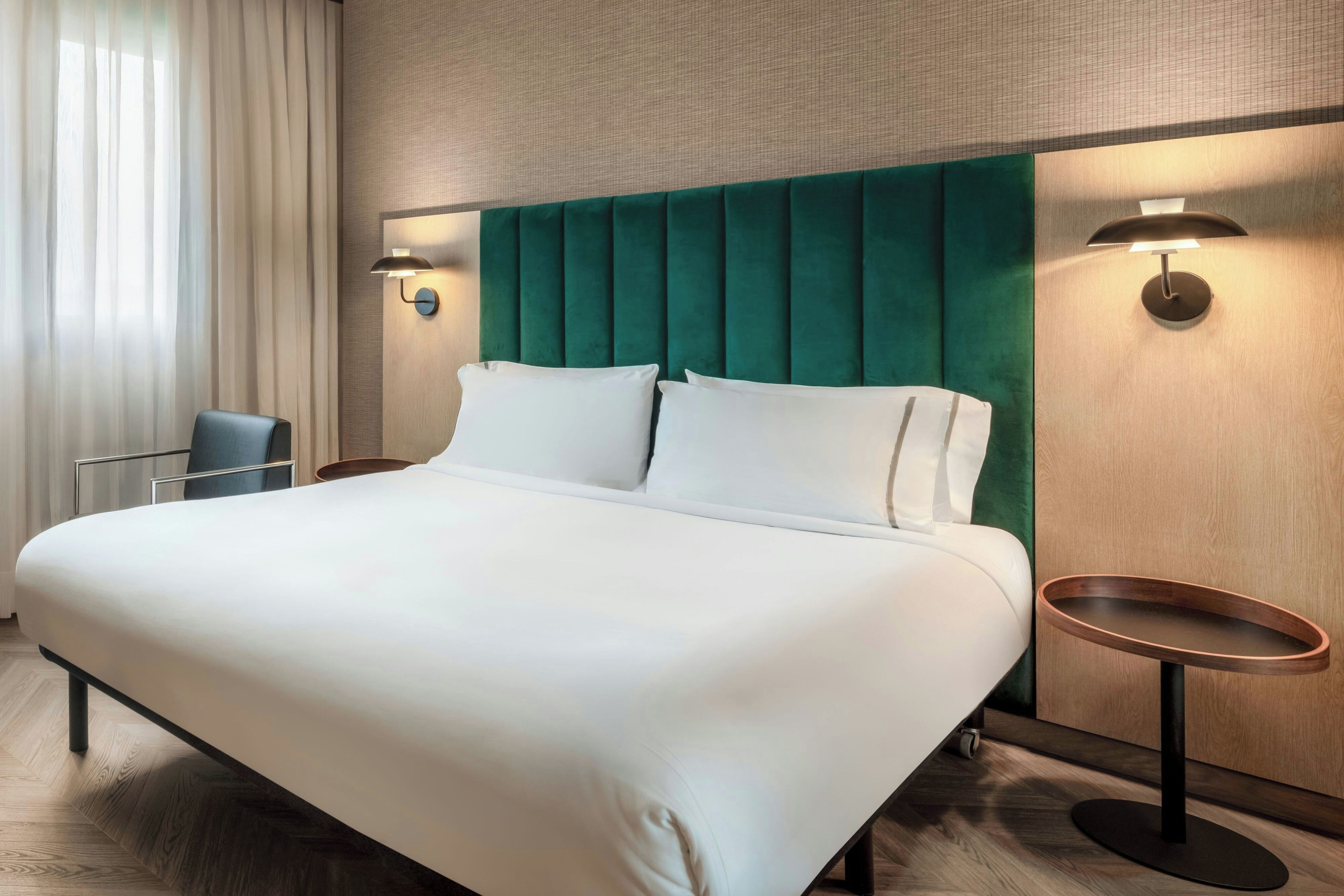 Una camera d'albergo con un letto grande, due comodini, una sedia e una finestra con tende.