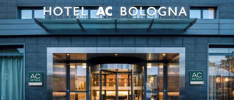 A modern hotel entrance with a revolving door, glass facade, and signage for 'HOTEL AC BOLOGNA'.