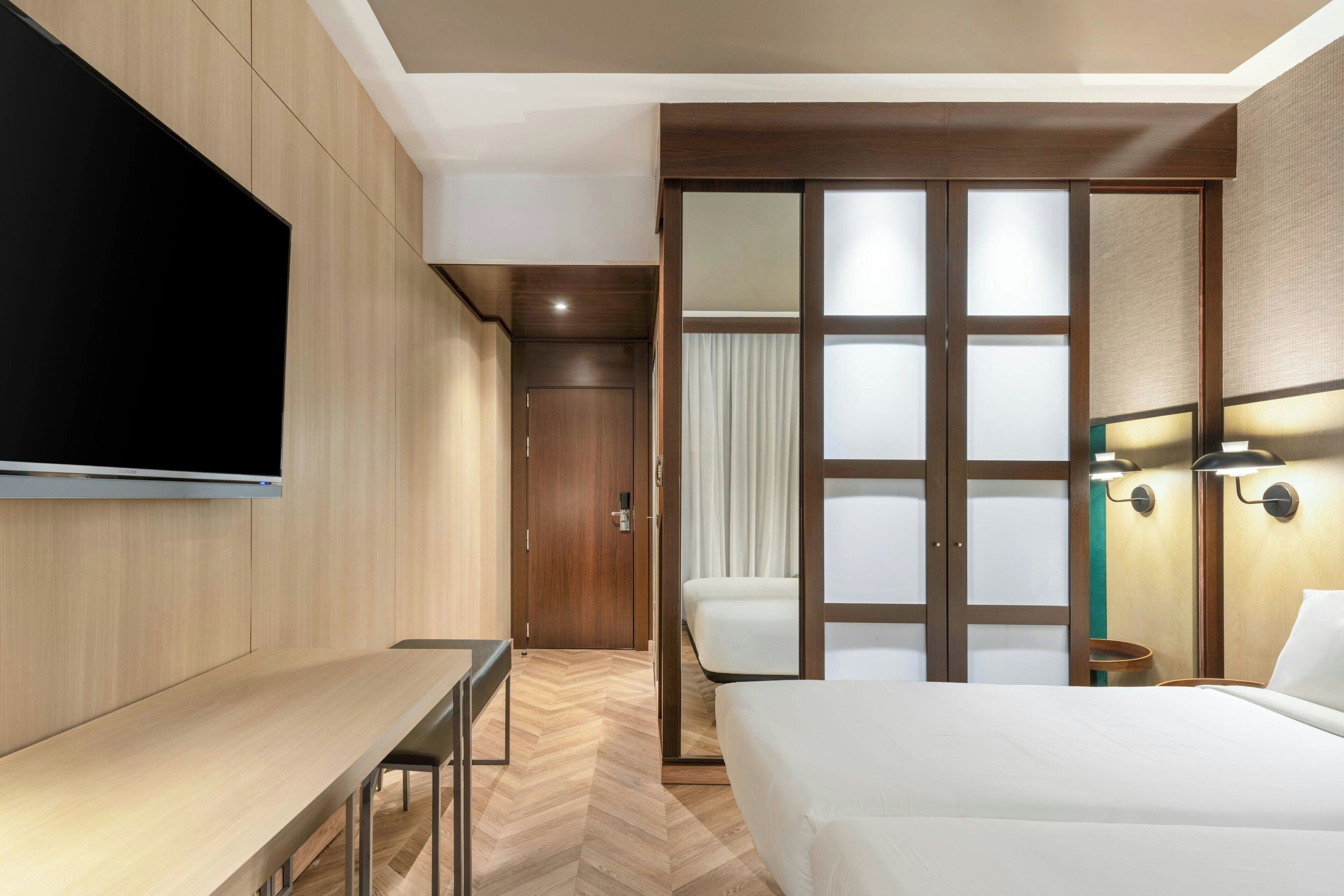 Une chambre d’hôtel équipée d’un lit, d’un bureau, d’une télévision et d’une porte coulissante donnant accès à la salle de bain.
