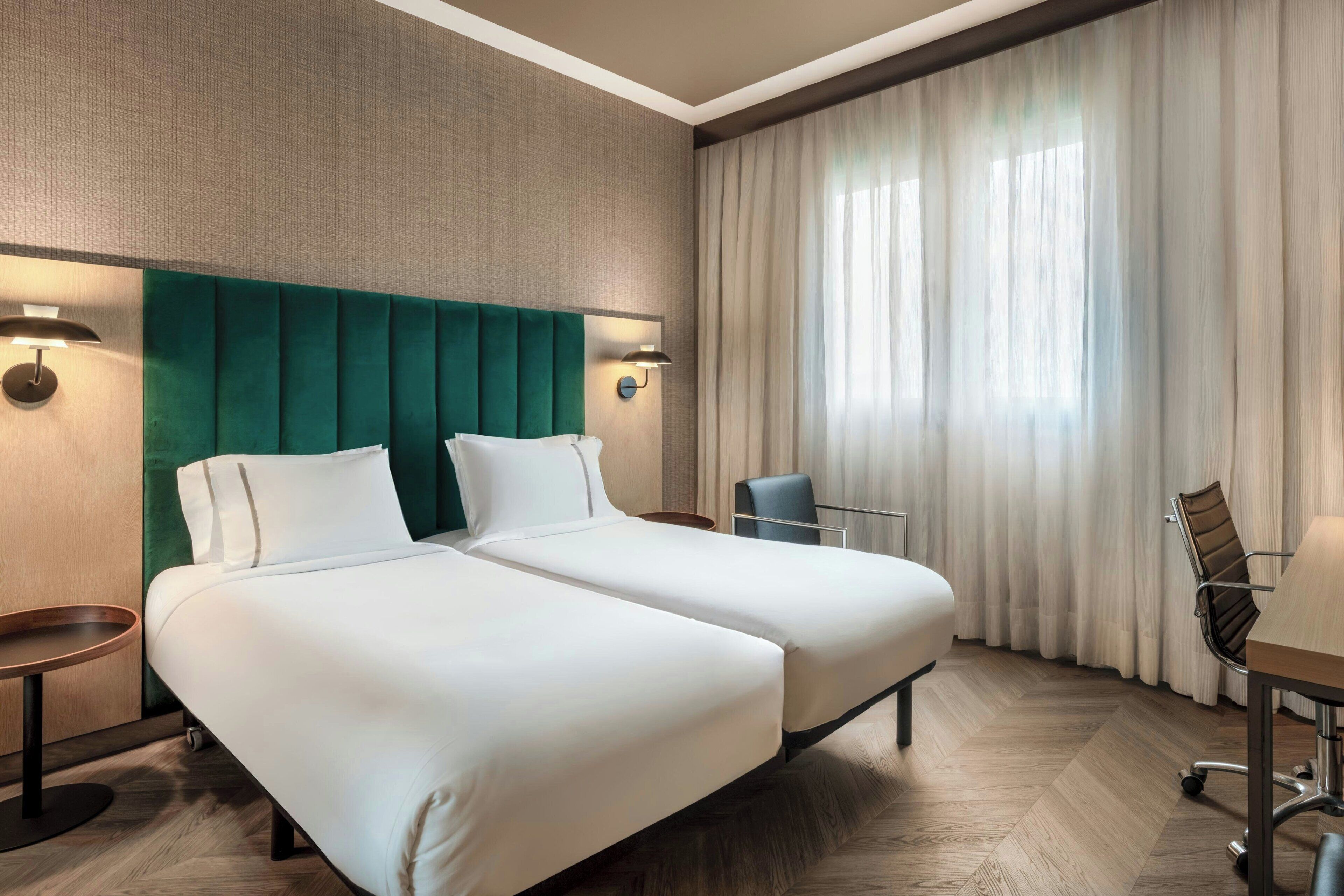 A hotel room with two beds, a desk, a chair, and a large window with curtains.