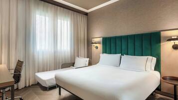 A hotel room with a large bed, a desk, a chair, and a window with curtains.
