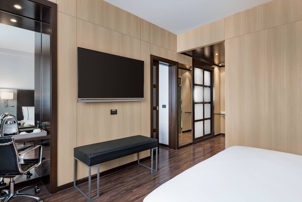A hotel room with a bed, a desk with a chair, a bench, a TV, and a door leading to another room. - AC Hotel Bologna by Marriott (Bologna)