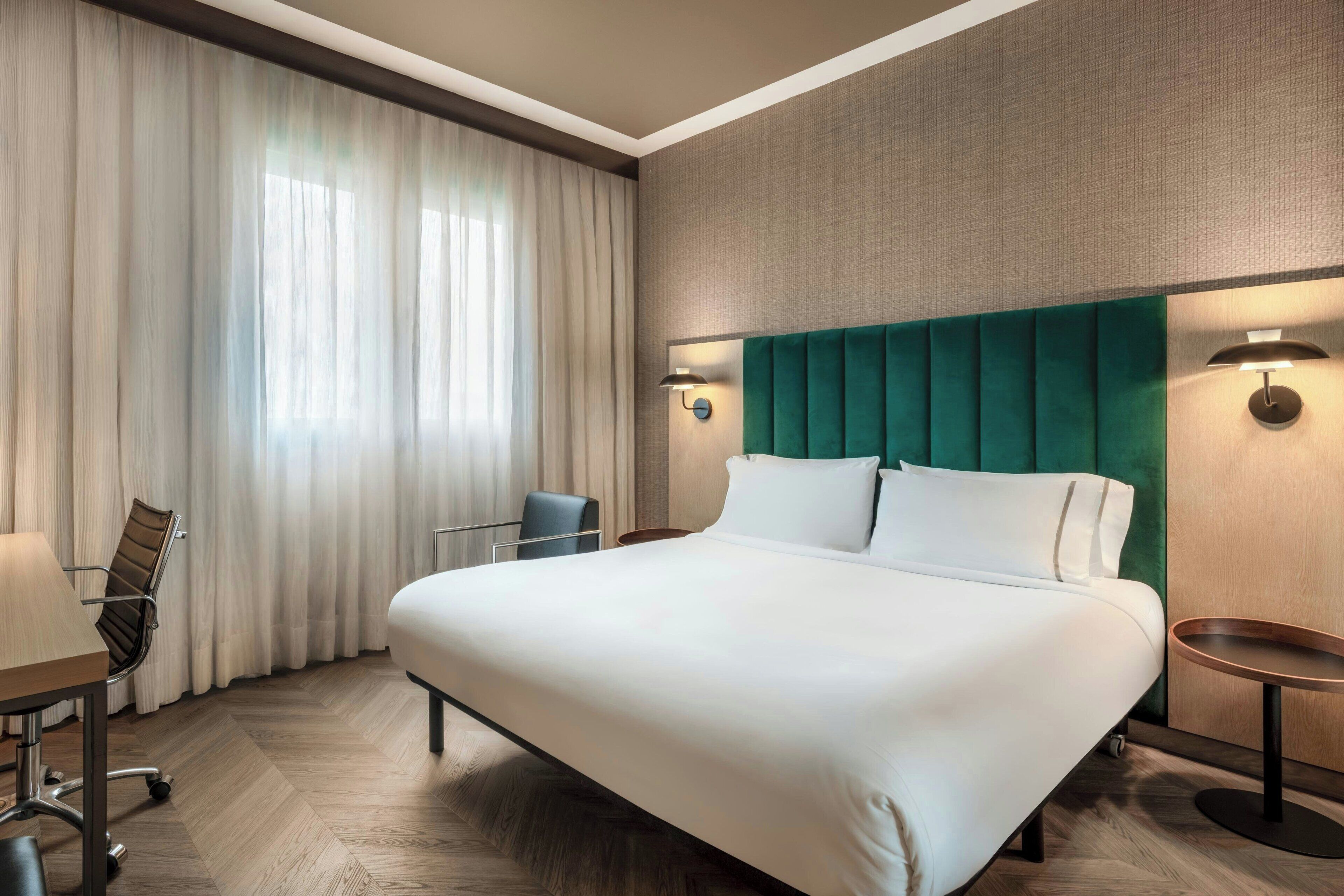 A hotel room with a large bed, a desk with a chair, a window with curtains, and a wall-mounted lamp.