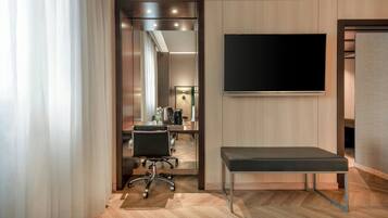 A modern hotel room with a wooden floor, a desk with a chair, a bench, a flat-screen TV, and a door leading to a bathroom.