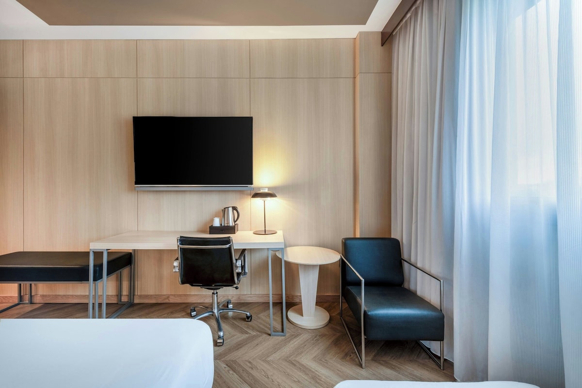 A hotel room with a bed, a desk with a chair, a television, and a window with curtains.