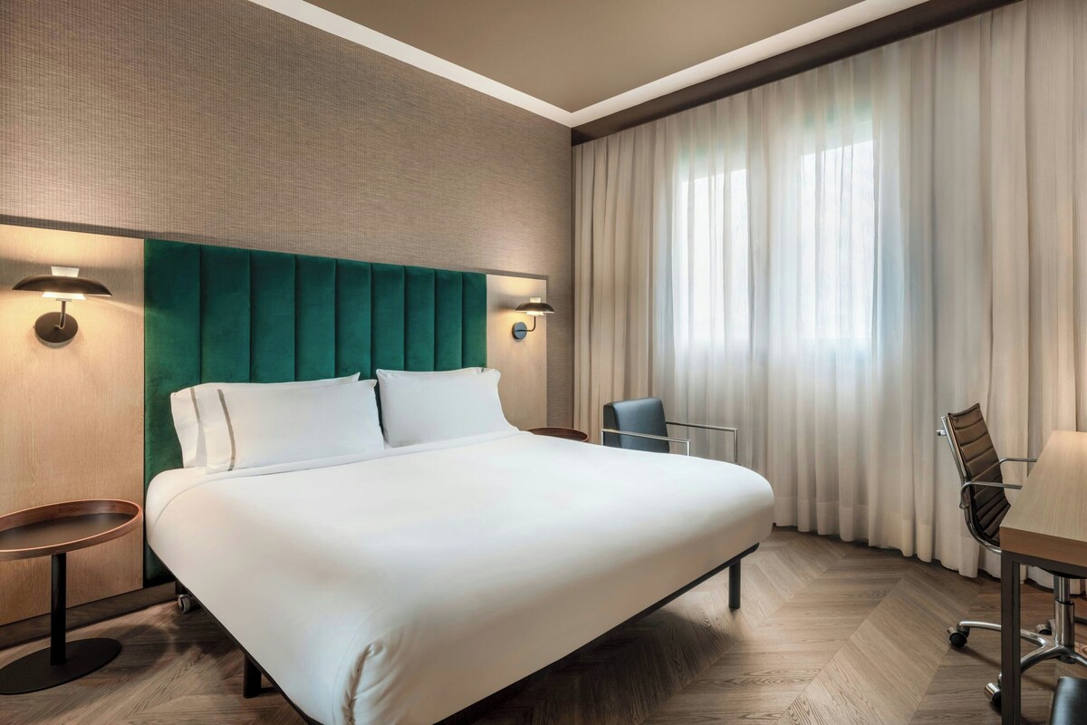 A hotel room with a large bed, two bedside tables, a desk with a chair, and a window with curtains.