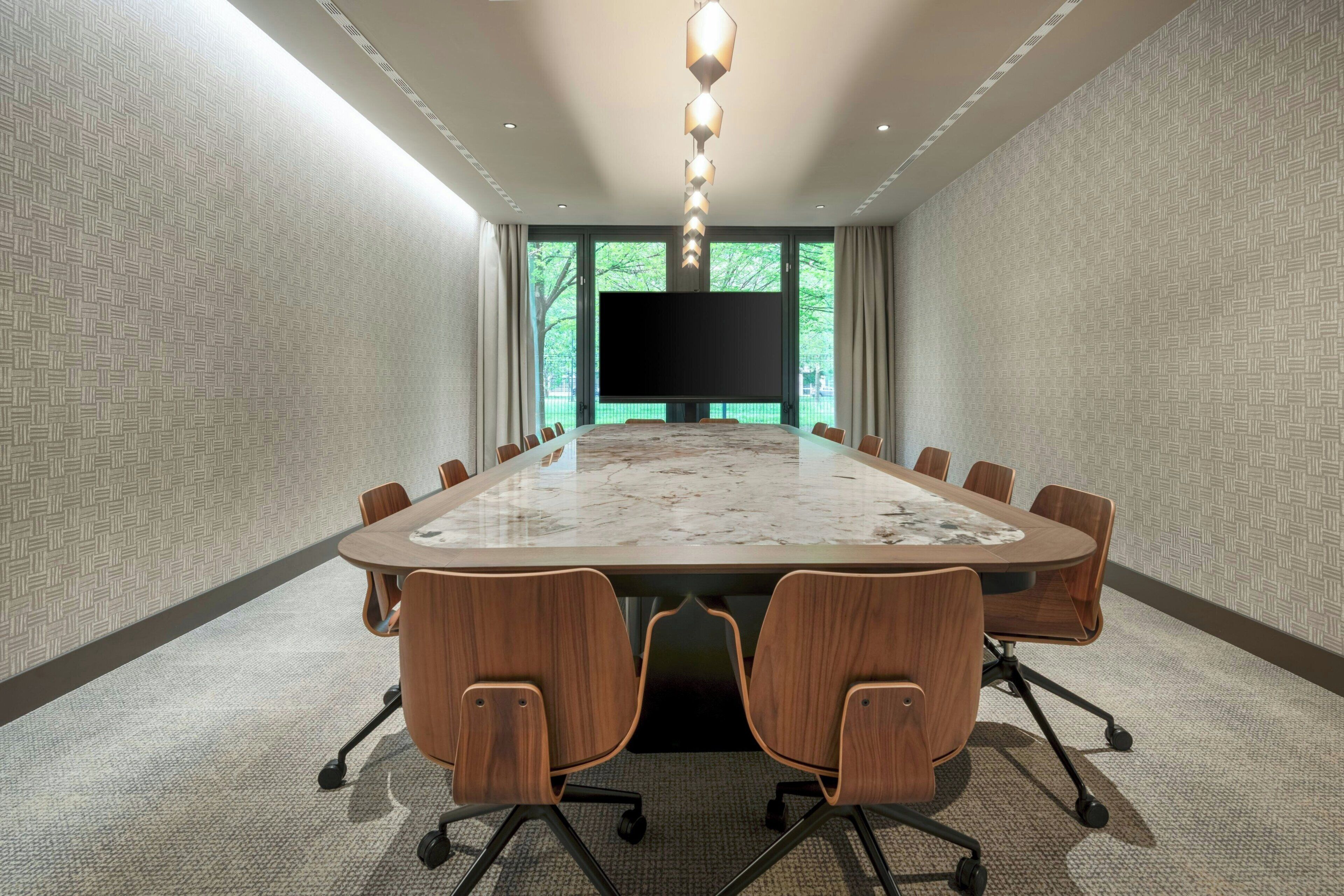A conference room with a long table, chairs, a large screen, and a view of greenery outside.