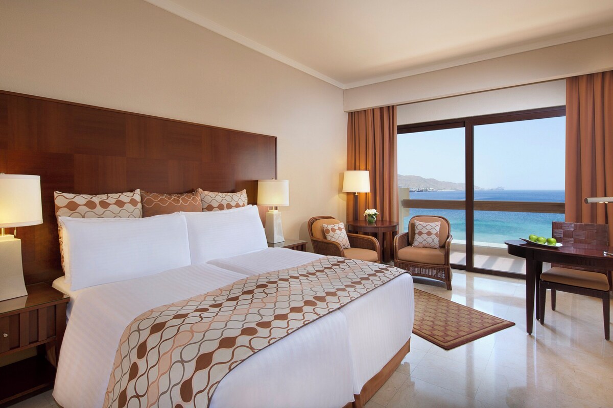 Une chambre d’hôtel avec un grand lit, deux tables de chevet avec des lampes, un bureau avec une chaise et une vue sur la mer.