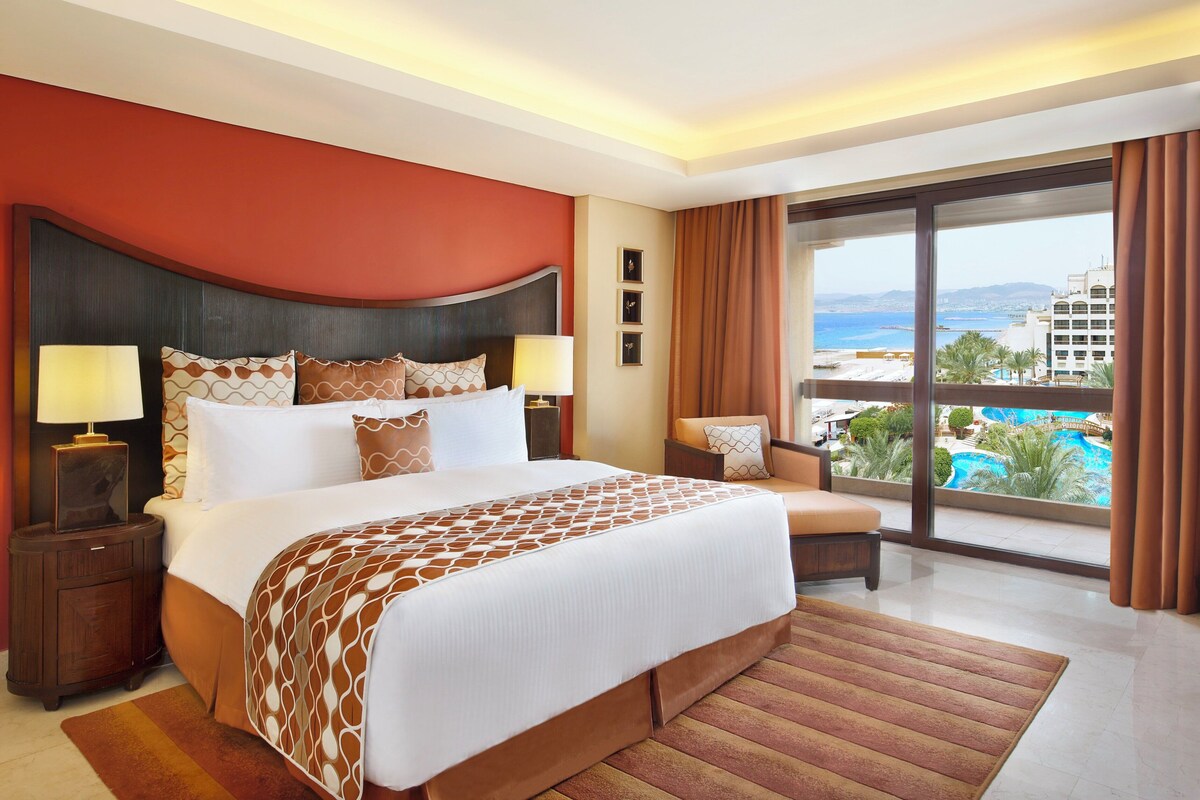 Une chambre d’hôtel avec un grand lit, deux tables de chevet avec des lampes, un canapé, une grande fenêtre donnant sur une plage et une piscine, et un mur d’accent rouge.