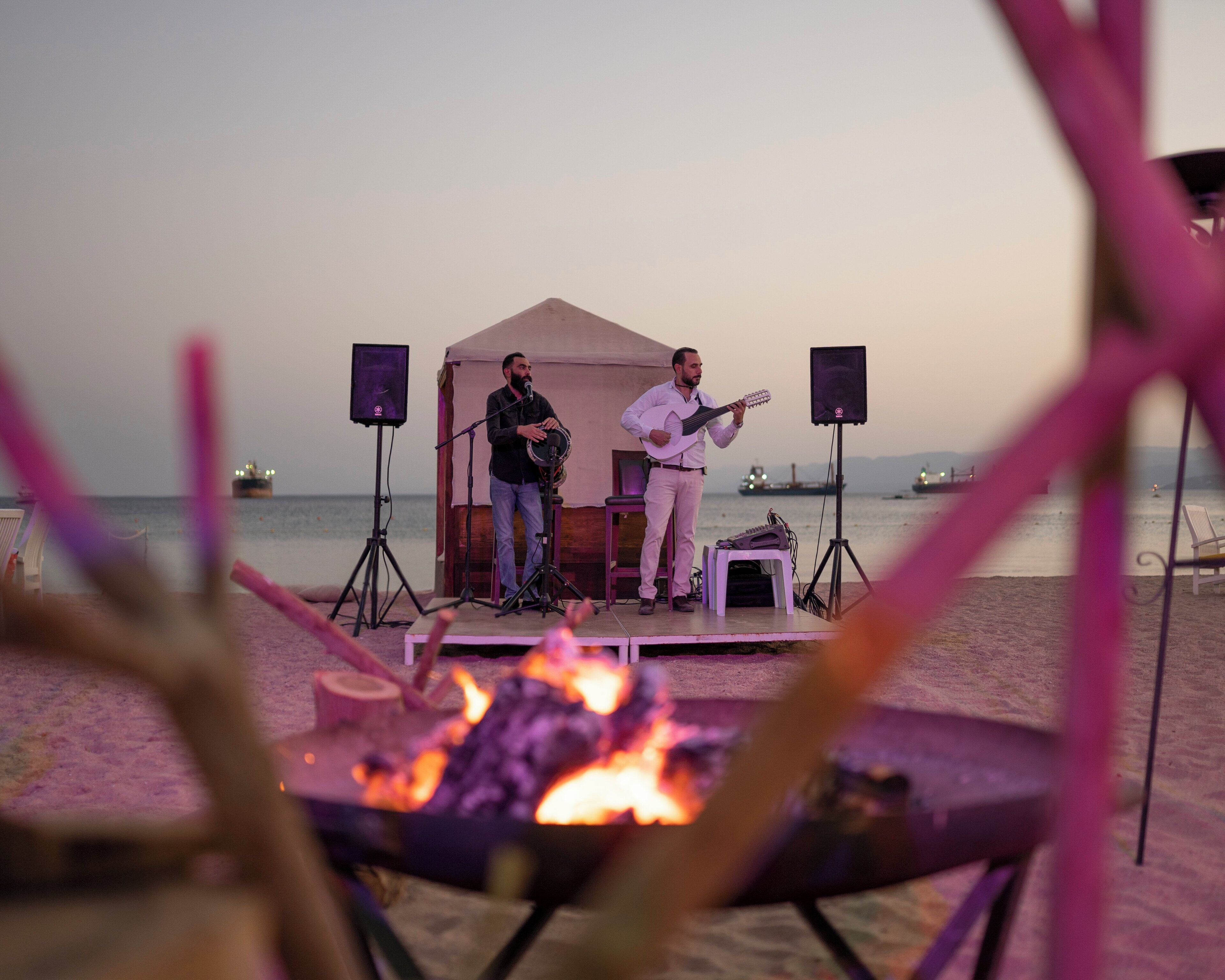 A stage with two musicians performing, a tent, and a fire pit on a beach.