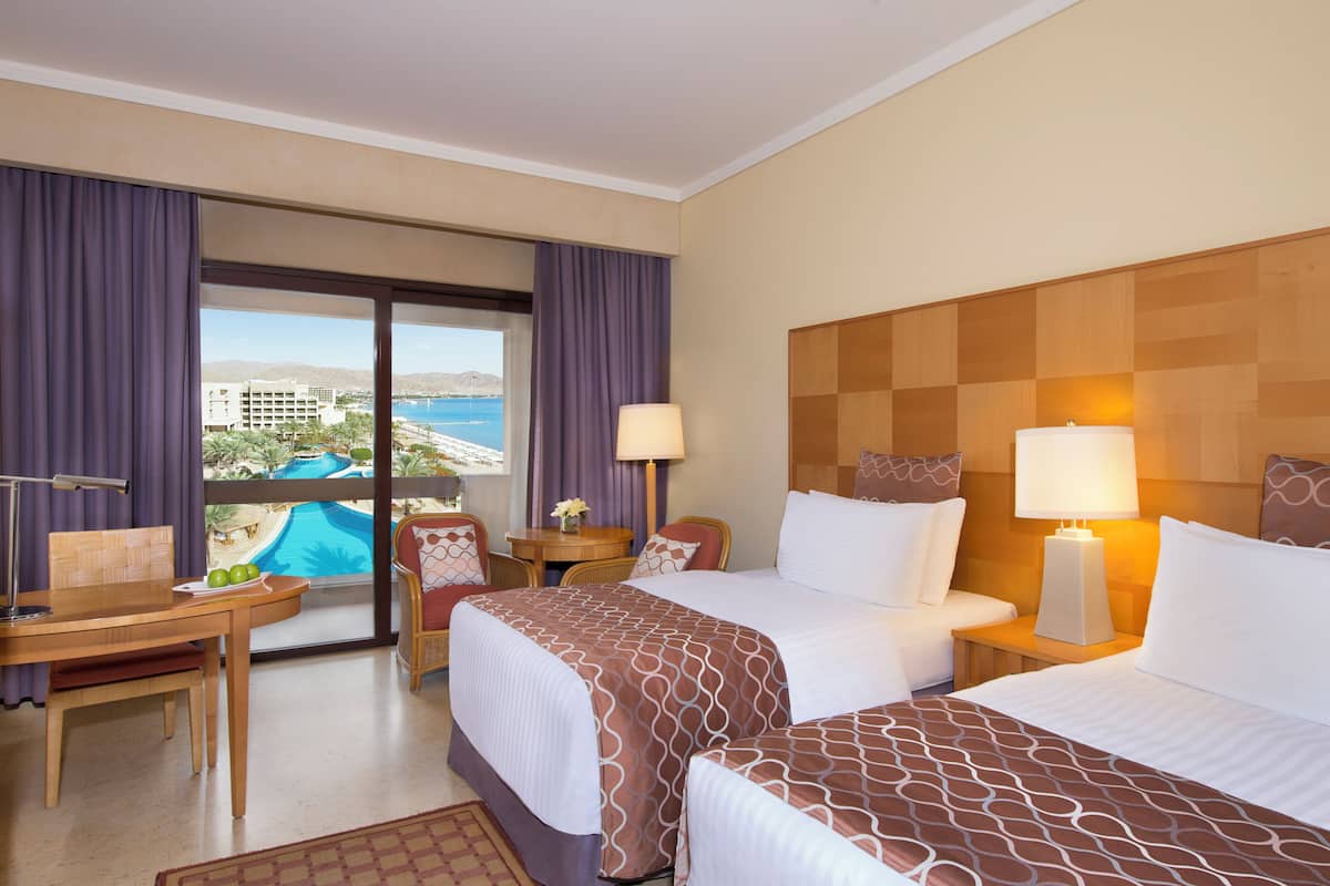 Ett hotellrum med två sängar, ett skrivbord och utsikt över en pool och byggnader.