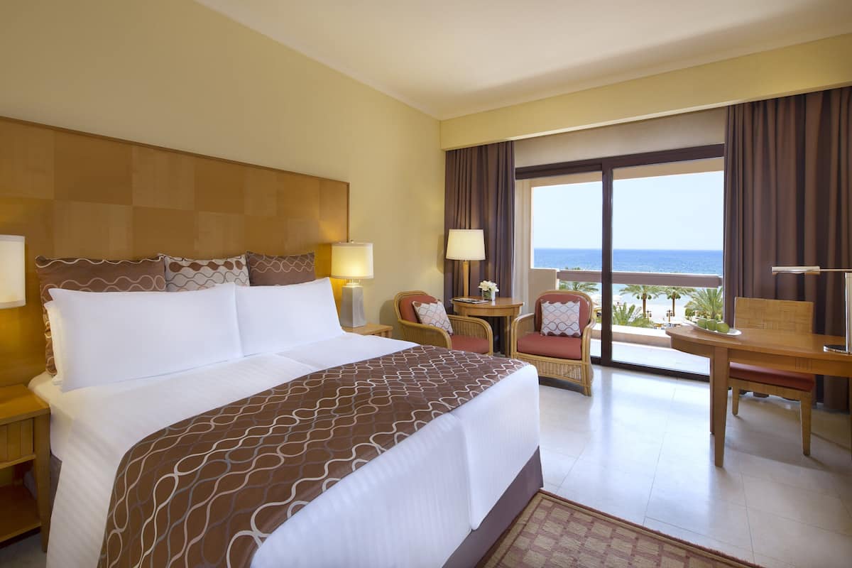 Habitación de hotel con una cama grande, dos mesitas de noche con lámparas, una silla, una pequeña mesa con lámpara y vista al mar a través de amplios ventanales.