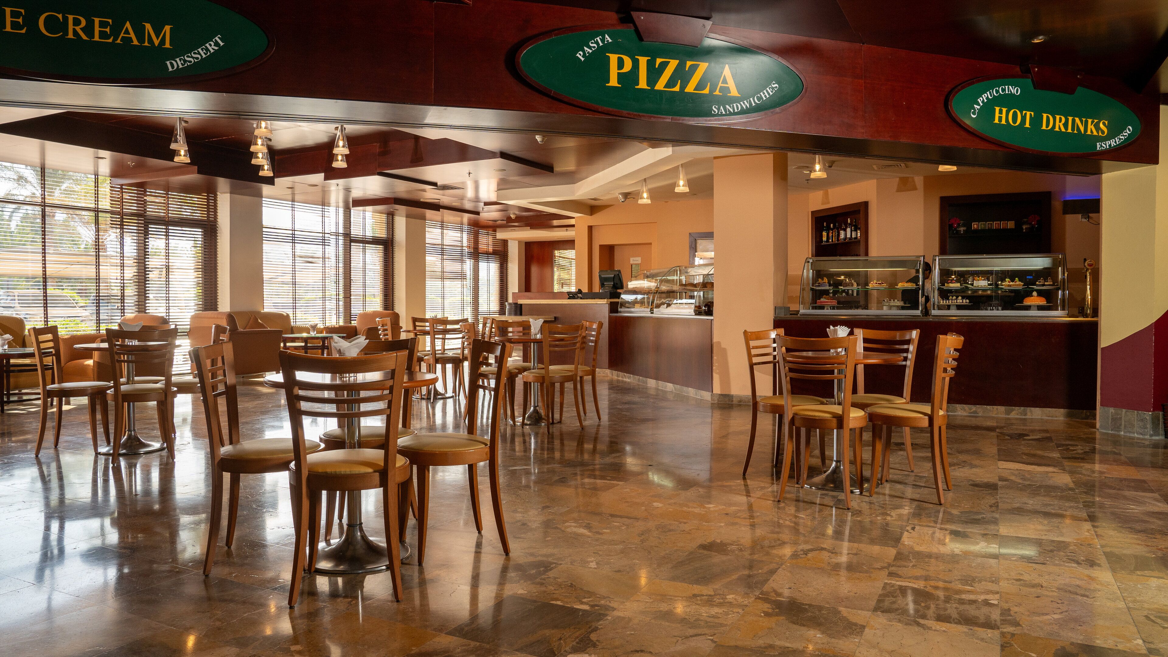 A restaurant interior with multiple seating areas, a counter with a display case, and signs indicating menu options like pizza and hot drinks.