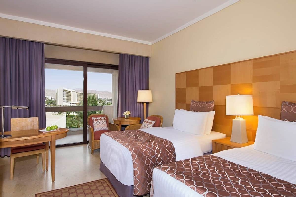 Ett hotellrum med två sängar, ett skrivbord, en stol, ett bord och utsikt över byggnader genom fönstret.
