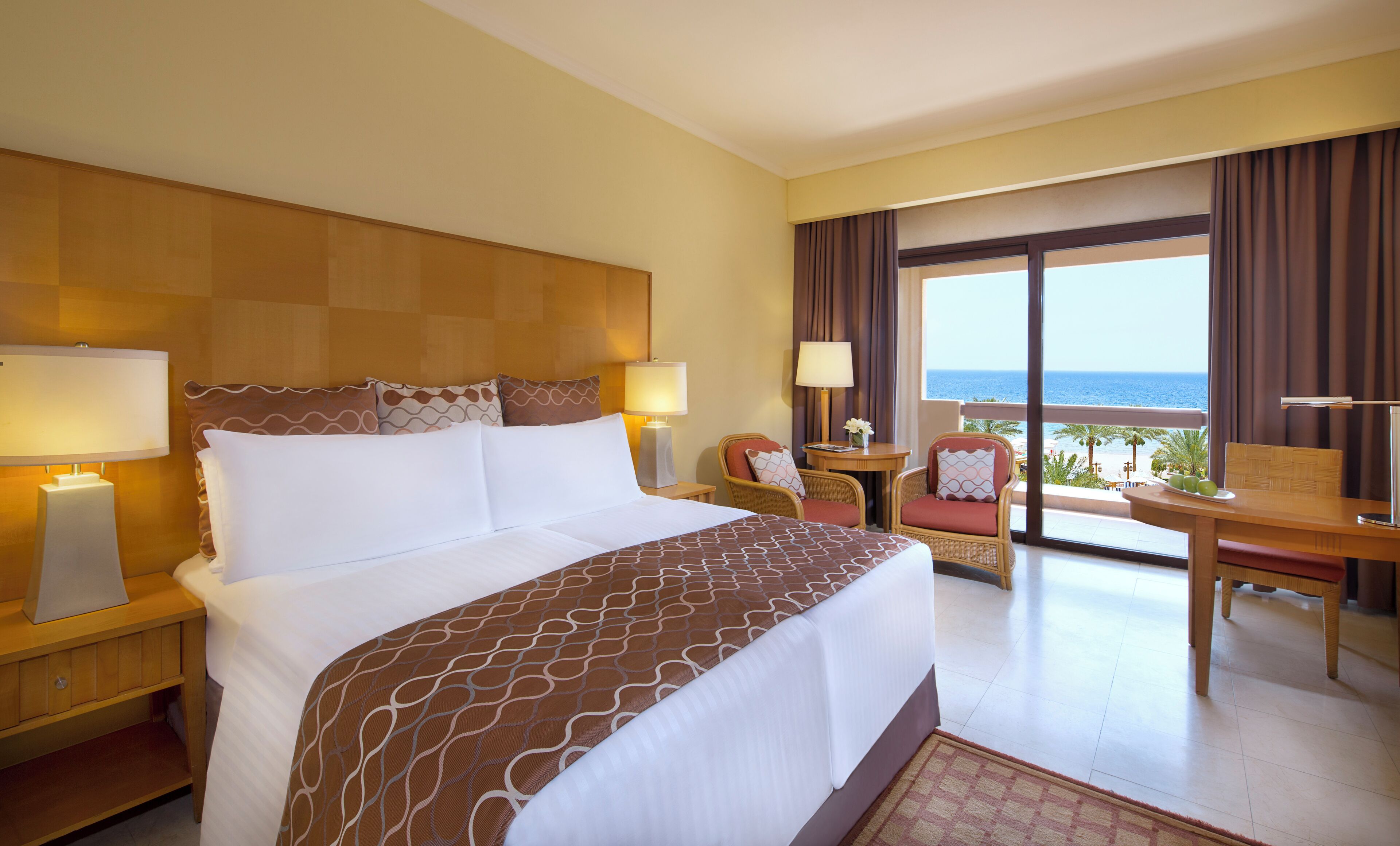 Habitación de hotel con una cama grande, dos mesitas de noche con lámparas, una pequeña mesa con una silla y un balcón con vista al mar.