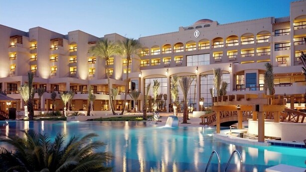 A hotel with a large swimming pool, multiple balconies, and palm trees.