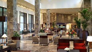 A spacious lobby with high ceilings, marble columns, and a chandelier. There are multiple seating areas with chairs and tables, and a reception desk in the background.
