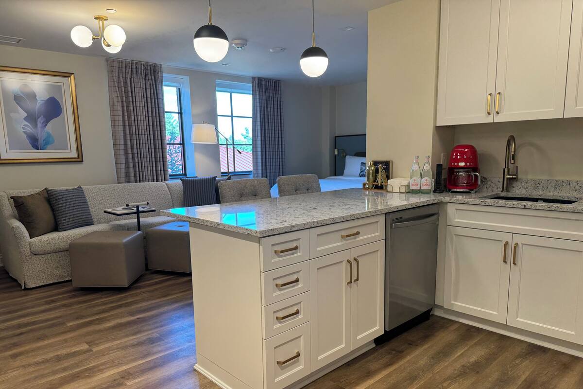 A modern kitchen with white cabinets, a granite countertop, and a built-in dishwasher. There is a dining area with a gray sofa and ottoman, and a large window with curtains.