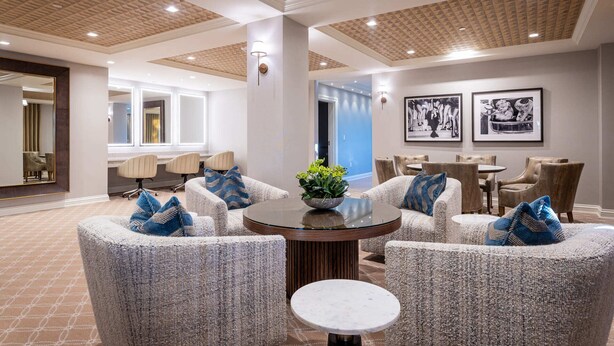 A modern hotel lobby with a round table surrounded by upholstered chairs, a large mirror, and framed artwork on the walls.