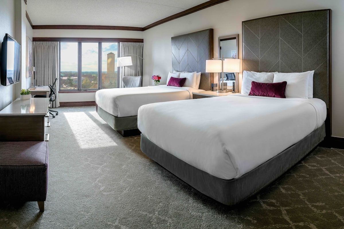 A hotel room with two beds, a desk, a chair, a TV, and a large window with a view.