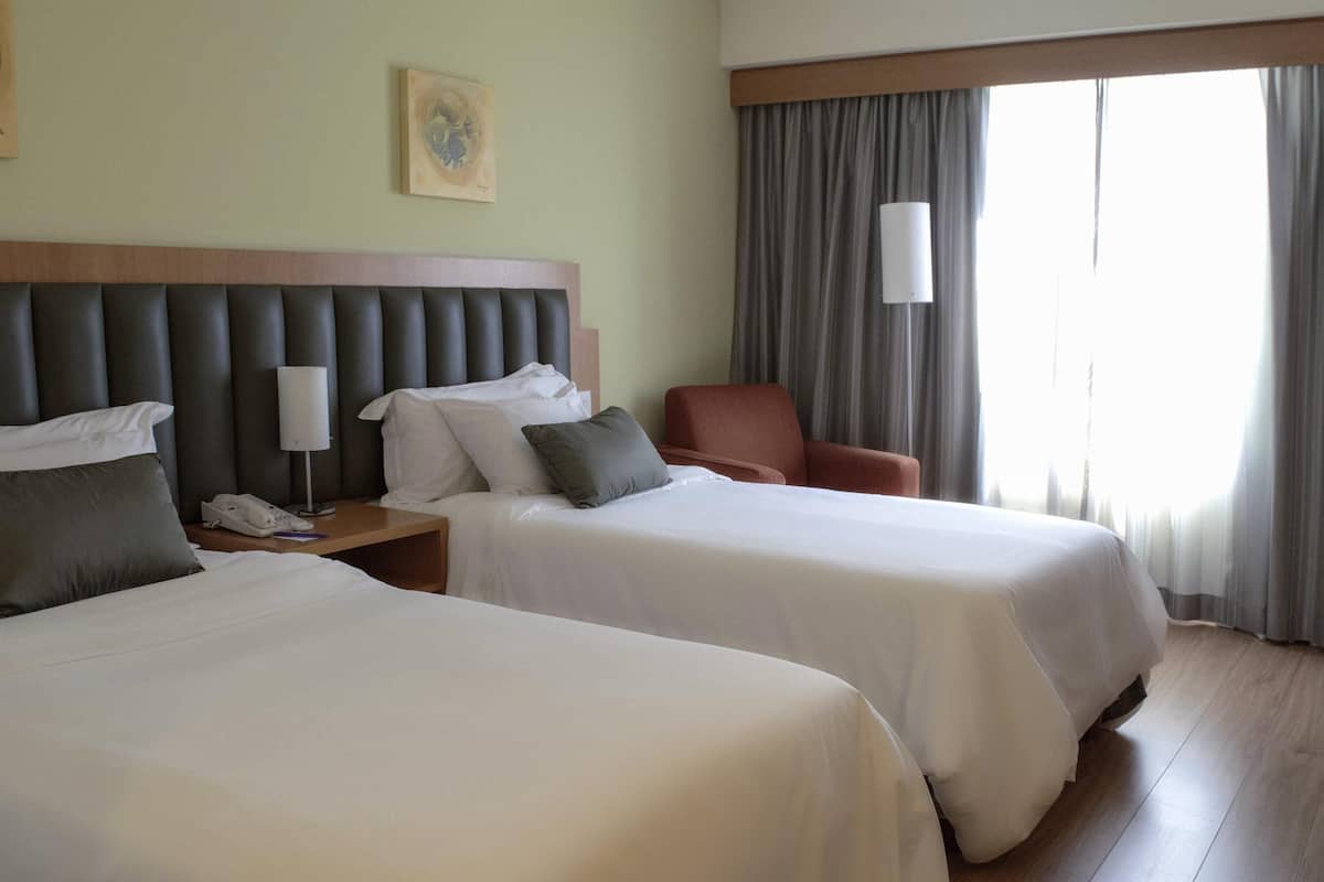 A hotel room with two beds, a wooden headboard, a nightstand with a phone, a chair, and a window with curtains.