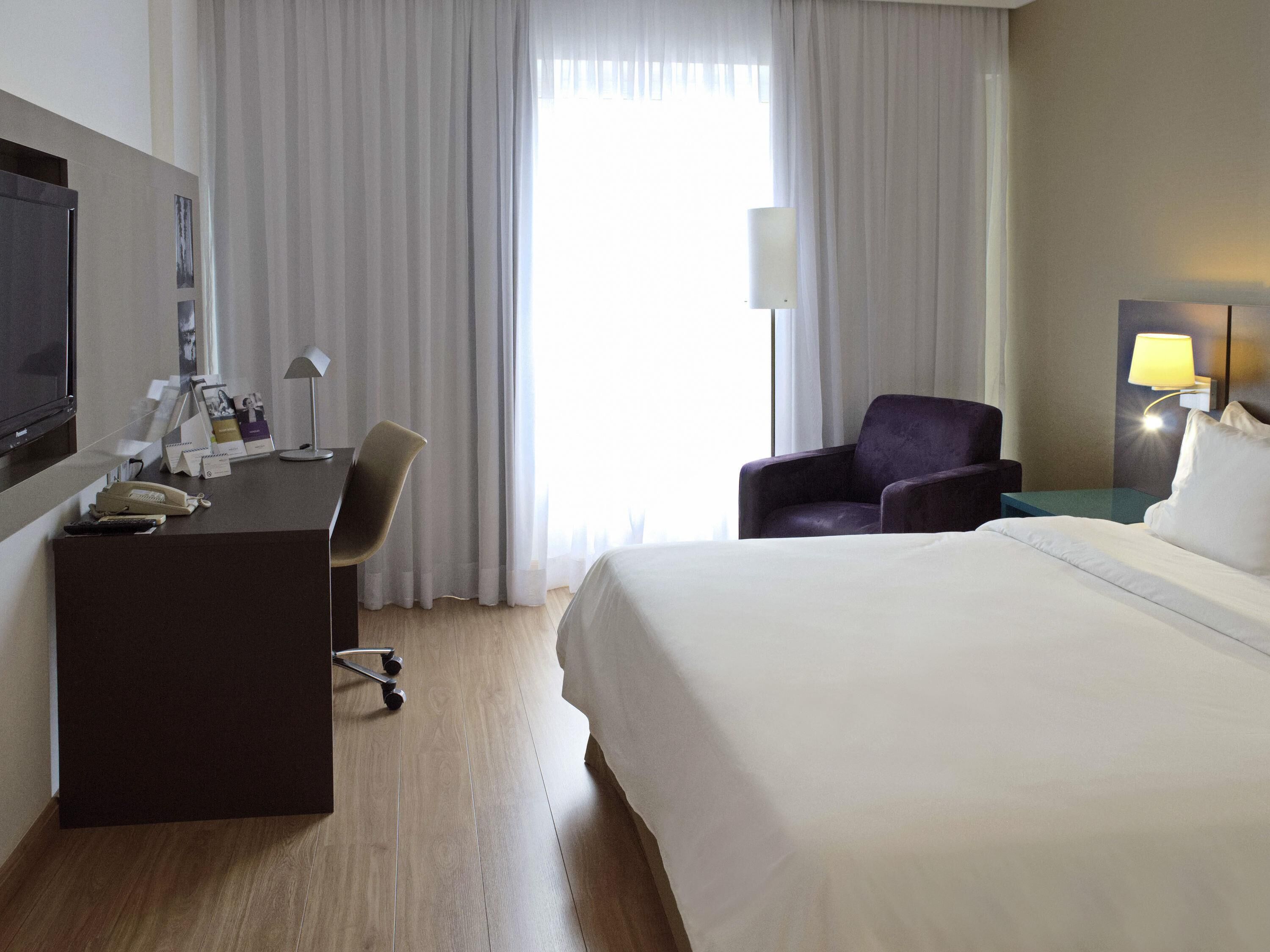 Quarto de hotel com uma cama, escrivaninha com cadeira, televisão, luminária e janela com cortinas.