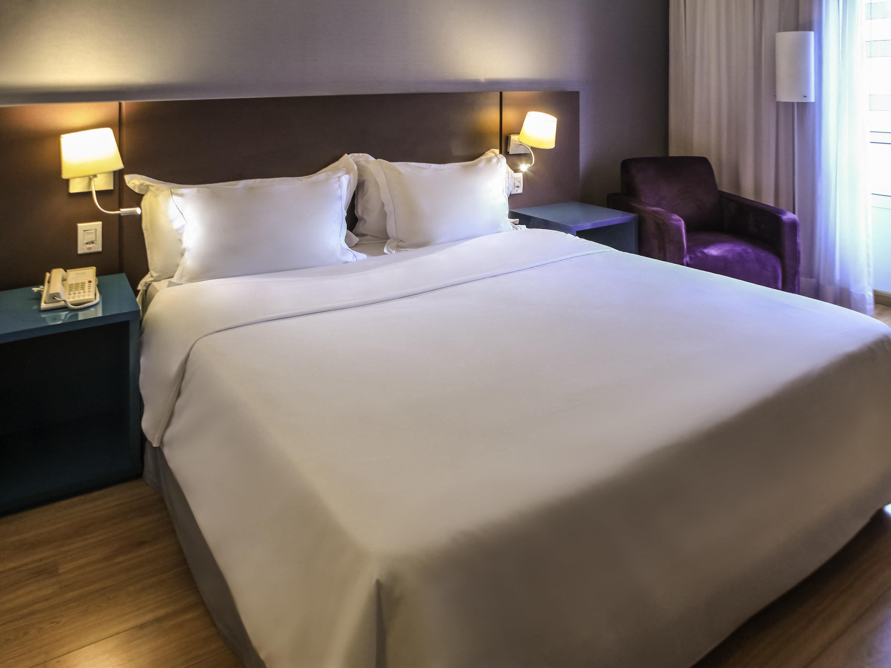Une chambre d’hôtel avec un grand lit, deux lampes de chevet, une table de chevet avec un téléphone et un fauteuil violet.