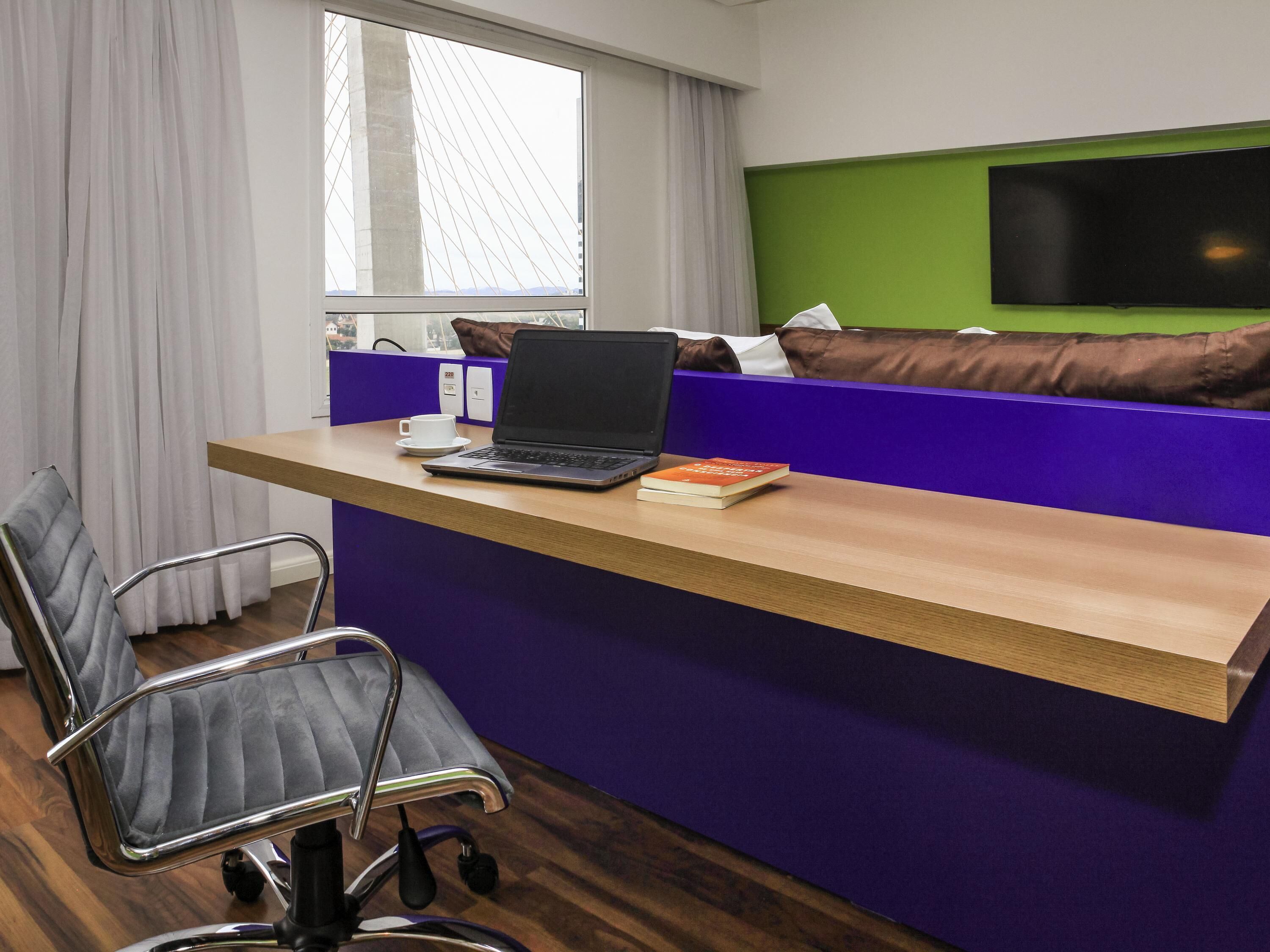 A modern office space with a desk, laptop, and a book. There is a window with a view of a bridge, a purple partition, and a green wall with a flat-screen TV.