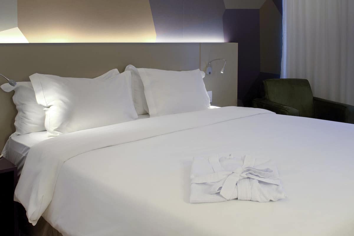 A neatly made bed with white linens and pillows, a folded towel on the bed, and a headboard with a light fixture.