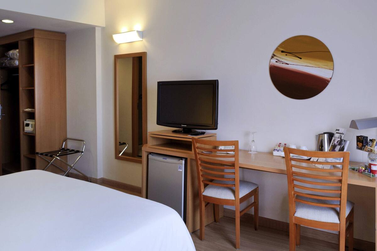A hotel room with a bed, a desk with a computer, two chairs, a refrigerator, a mirror, and a wall-mounted picture.