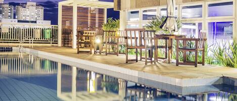 Zona de piscina con mobiliario de madera y vistas del horizonte urbano al caer la noche.