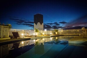 A nighttime pool area with a modern building in the background, lounge chairs, and a fence.