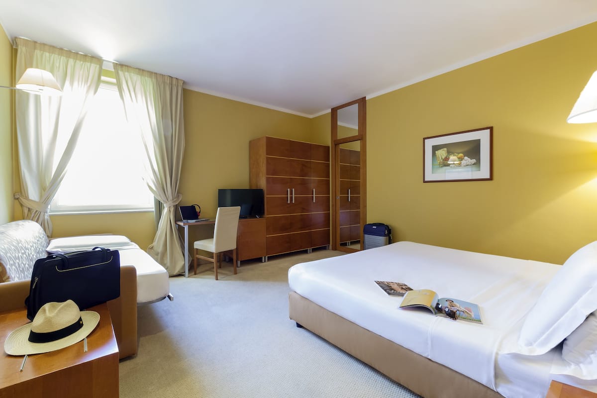 A hotel room with a bed, a desk with a chair, a wardrobe, and a window with curtains.